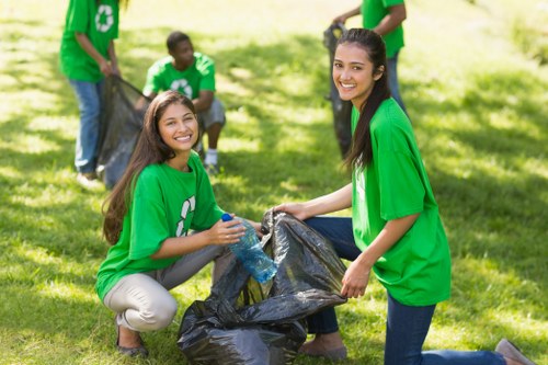 A group of young volunteers participating in a community clean-up event outdoors on a bright, sunny day. In the foreground, two women are kneeling on the grass, smiling at the camera while holding a large black rubbish bag filled with collected waste; one woman is wearing a green T-shirt, beige trousers, and has long brown hair, while the other is also in a green T-shirt, dark jeans, and has long dark hair. In the background, three additional volunteers are seen contributing to the rubbish collection, also dressed in green T-shirts, with some holding black rubbish bags and others actively gathering litter. The scene is set on a lush, green lawn with a backdrop of trees and natural light filtering through the leaves, creating a bright and lively atmosphere. This image illustrates community efforts in waste management and rubbish removal, aligning with services offered by companies such as Office Clearance Balham in the local area around Balham or postcode SW12.