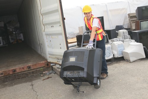 A worker wearing a yellow safety helmet, a high-visibility vest with red and yellow stripes, and grey gloves is standing outdoors on a paved surface, lifting and moving an old computer monitor and a black electronic waste item. The background shows a partially open shipping container, with various discarded electronics and appliances packed nearby, indicating a rubbish removal or clearance activity related to office or electronic waste disposal in Balham. The area appears to be part of a commercial or industrial site, with the container on the left side and some wooden pallets and additional electronic items stacked against an outdoor wall or fence. Natural daylight illuminates the scene, emphasizing the textures and colors of the electronic waste and the workerâ€™s safety gear. Office Clearance Balham's services are relevant to this environmentally responsible rubbish collection, supporting proper disposal of electronics from local offices or facilities in the Balham area.