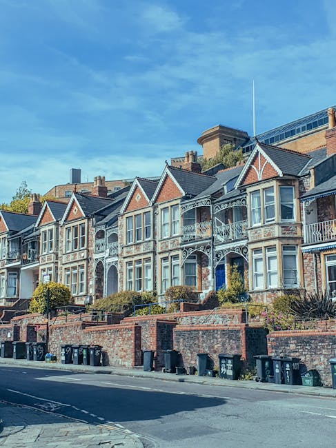 A row of historic bay-fronted Victorian terraced houses with ornate white ironwork balconies and decorative brickwork in shades of red and cream, situated on a sloped street. In the foreground, there 