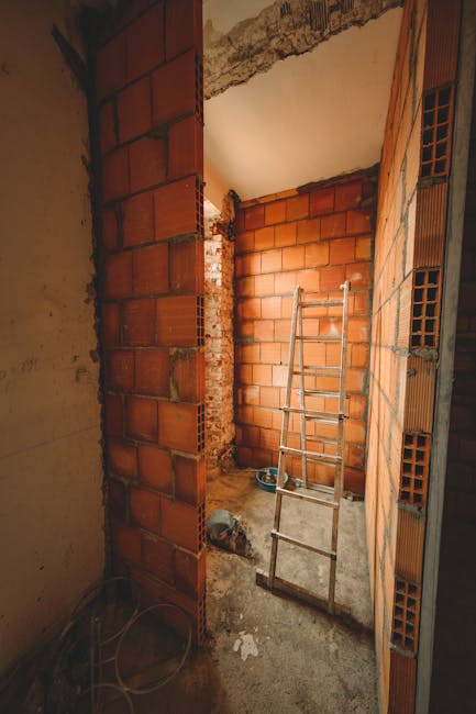 The image depicts a cluttered storage area with a rough brick wall background illuminated by natural light from a window on the right. In the foreground, there is a stack of nine cardboard boxes label