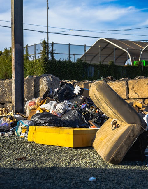 An outdoor scene showing a collection of mixed domestic and recyclable waste piled on the edge of a paved area. The refuse includes black plastic garbage bags, a yellow plastic tray, a rolled-up rubbe