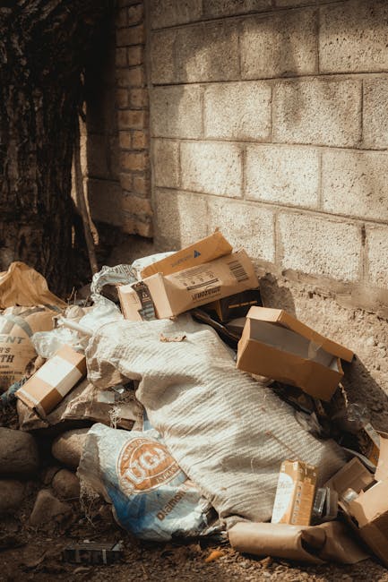 An outdoor scene showing a collection of mixed domestic and recyclable waste piled on the edge of a paved area. The refuse includes black plastic garbage bags, a yellow plastic tray, a rolled-up rubbe