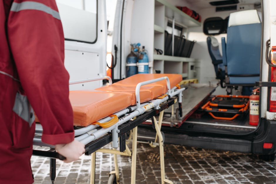 The image shows the interior of a commercial emergency response vehicle, with a person dressed in a red uniform loading an orange, padded hospital stretcher onto a frame at the rear of the vehicle. Th